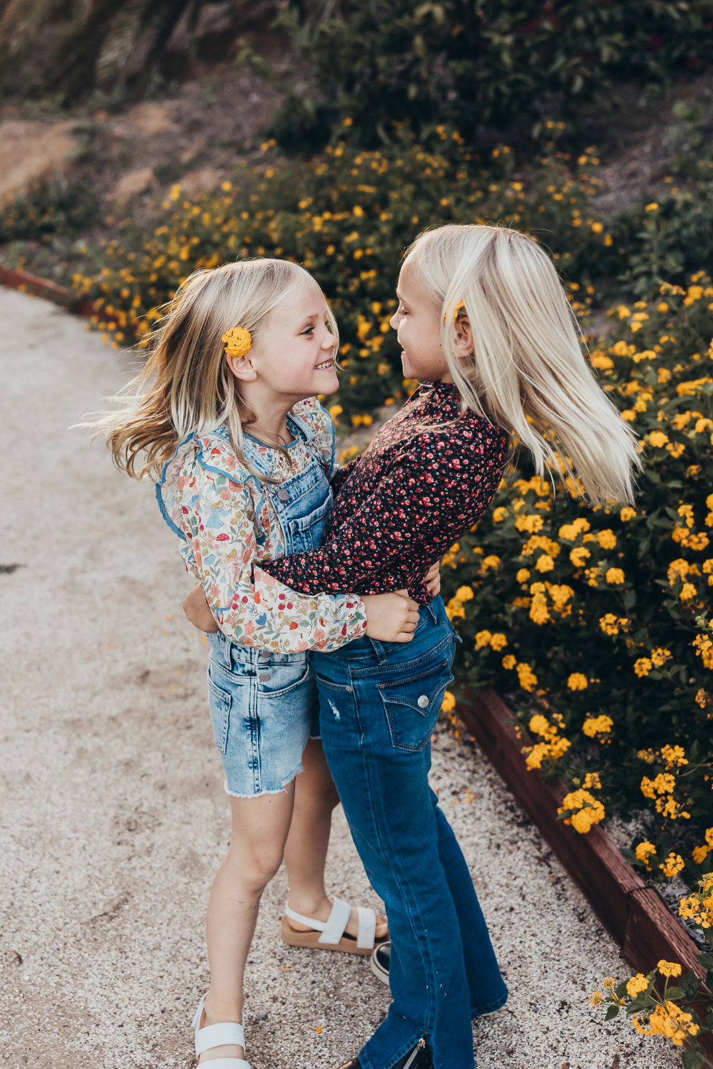 Two young sisters hug each other while smiling beside bright yellow flowers in a garden. Their joyful expressions, windblown hair, and sweet embrace capture the close bond between siblings in a relaxed outdoor family portrait.