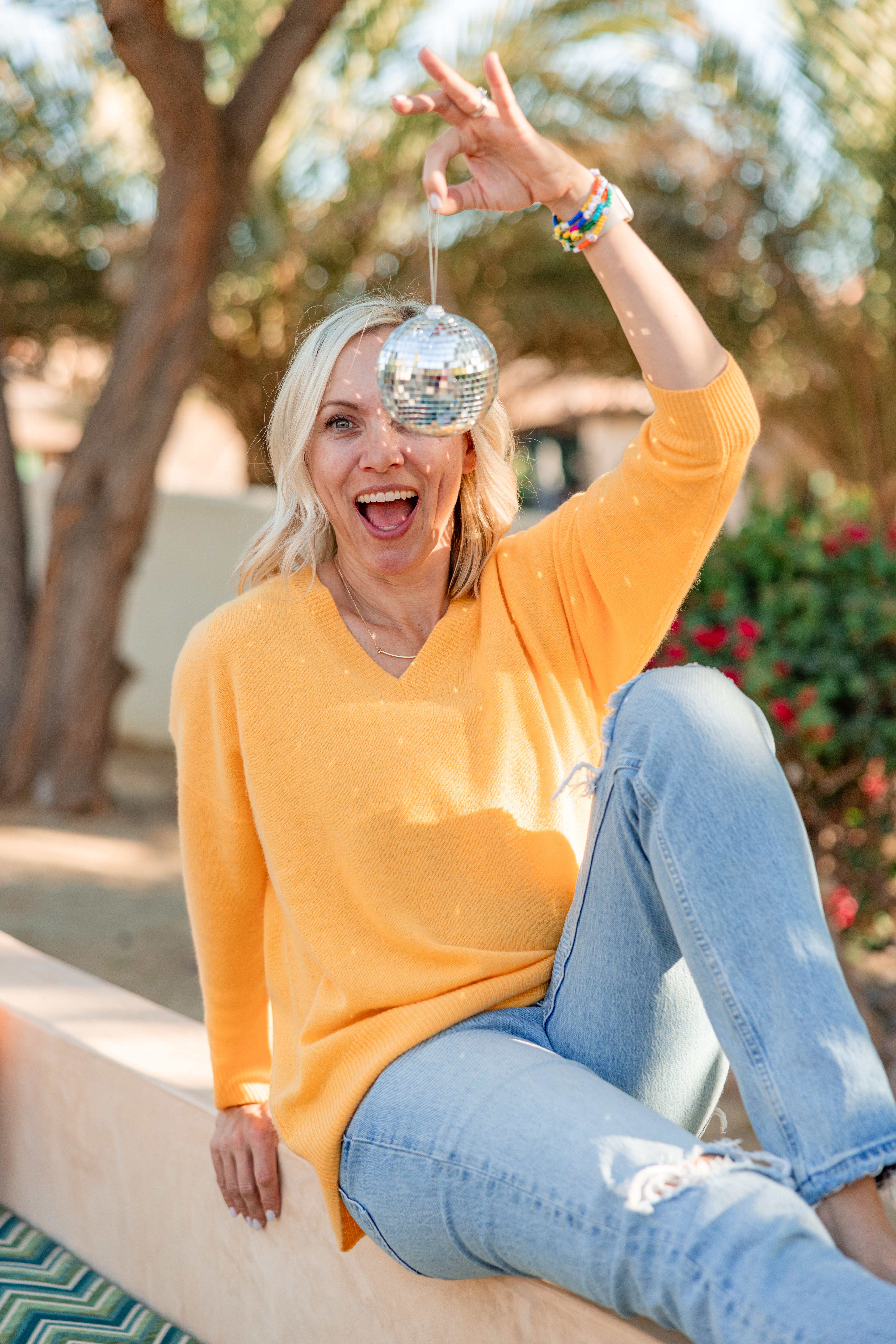 Photography mentor Christine Dammann sits outdoors in a yellow sweater and jeans, smiling while holding a small disco ball in the sunlight, reflecting the relaxed and playful personality she brings to photography education and mentoring.