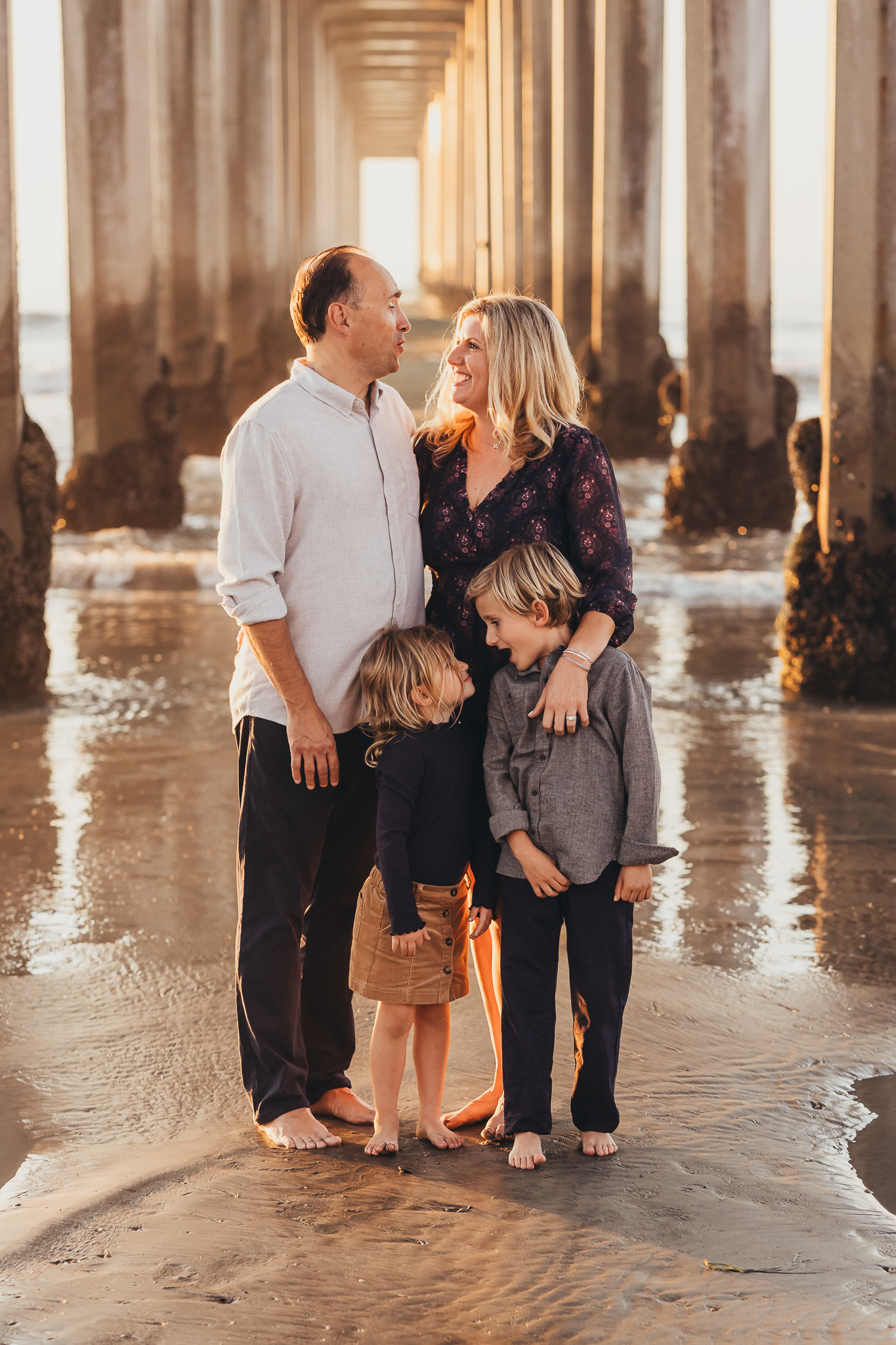 A family of four stands together beneath a pier at the beach during golden hour, laughing and connecting as warm sunlight fills the background, showing the natural movement and interaction at the heart of a relaxed family photography session.