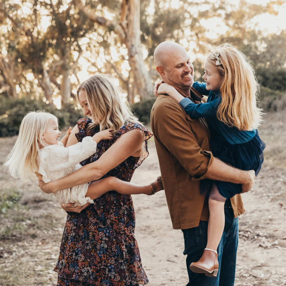 Family of four playing together on a wooded trail in warm evening light, with each parent holding one of their young daughters. A playful family photo filled with movement, laughter, and natural interaction.