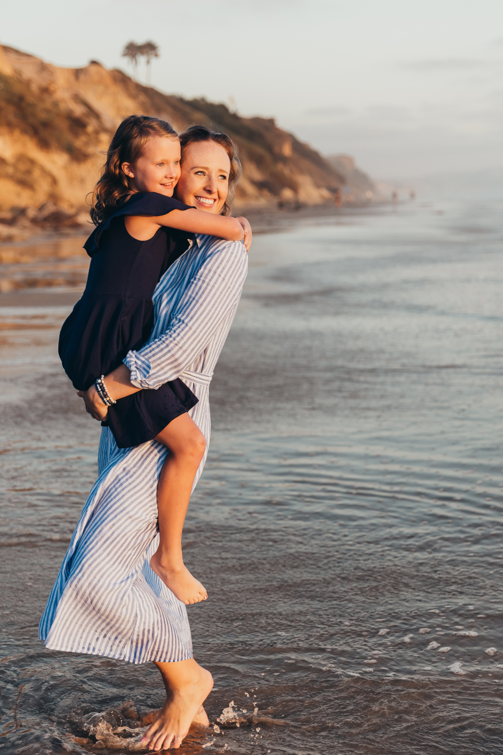 Mother carrying her young daughter through shallow ocean water during golden hour, both smiling as warm sunlight hits the coastal cliffs behind them. A sweet place for families to take pictures in San Diego.